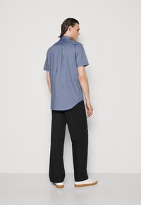 Mann, der von hinten zu sehen ist, trägt ein blaues kurzärmeliges Hemd mit Knöpfen, schwarze weite Hose und weiße Sneakers mit Gummisohle.