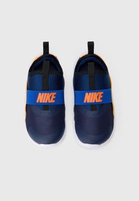 Para granatowych butów Nike typu slip-on z pomarańczowym logo na niebieskim pasku i białymi podeszwami, widziane z góry na białym tle.