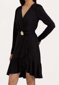 Svart wrap-klänning med långa ärmar, volangkant och en gyllene spänne. Tillverkad av mjukt tyg, med en djup V-ringning.