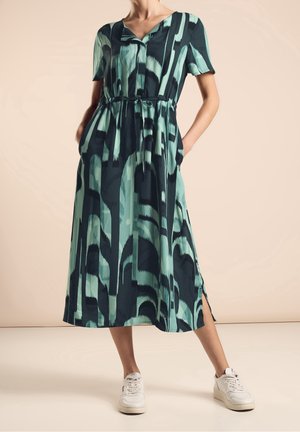 Maxikleid in Petrol und Marineblau mit abstraktem Muster, kurzen Ärmeln, Gürtel in der Taille, seitlichen Taschen und seitlichen Schlitzen; kombiniert mit weißen Sneakern.