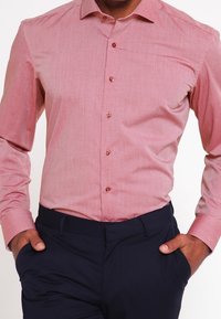 Chemise rose clair à manches longues en tissu lisse, avec un col boutonné et des boutons marron, associée à un pantalon bleu marine.