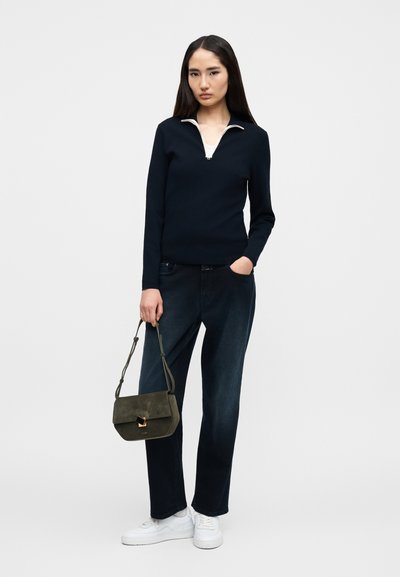 Pull bleu marine à manches longues avec une fermeture éclair blanche, jean en denim foncé, baskets blanches, et sac à main en daim vert avec un accent doré.