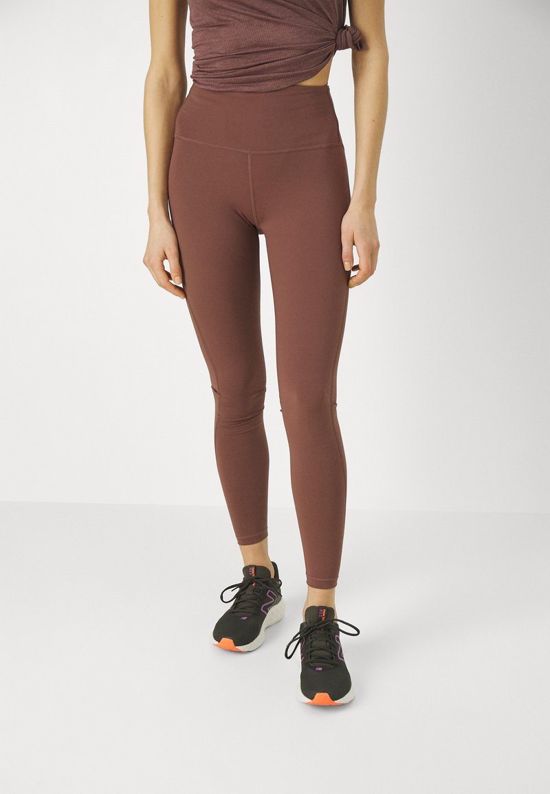 Leggings marron à taille haute, côtelés, avec une texture lisse et une coupe près du corps, associés à des chaussures de sport noires avec des accents oranges.