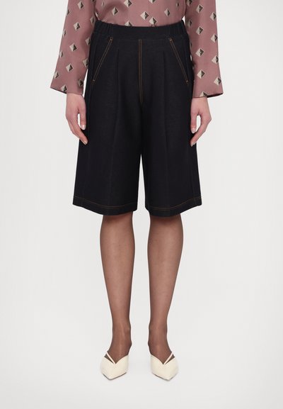 Shorts neri a pieghe con cuciture arancioni visibili, realizzati in un materiale texturizzato. Indossati con scarpe a punta color crema.