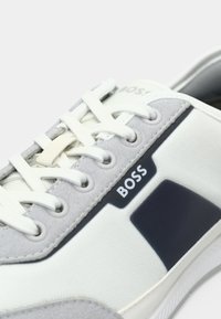 Biało-szary sneaker z cholewką wykonaną z tkaniny i zamszu, kontrastującymi granatowymi akcentami, okrągłymi sznurowadłami oraz naszywką z logo po boku.