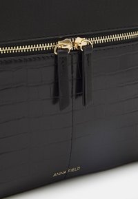 Musta tekonahkakassi, jossa on krokotiilinkuos, kultaiset vetoketjut ja yksityiskohdat, sekä brändin logo kohokuvioituna edessä.