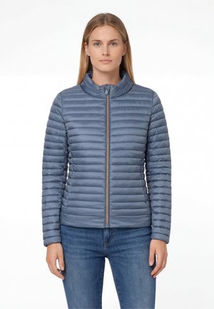 Donna che indossa una giacca imbottita trapuntata azzurro chiaro chiusa con la cerniera, abbinata a jeans blu, in piedi contro uno sfondo bianco.