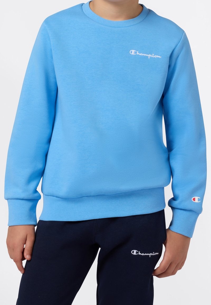 Enfant portant un sweat-shirt Champion bleu clair et un pantalon de jogging Champion bleu marine, debout les bras détendus le long du corps.