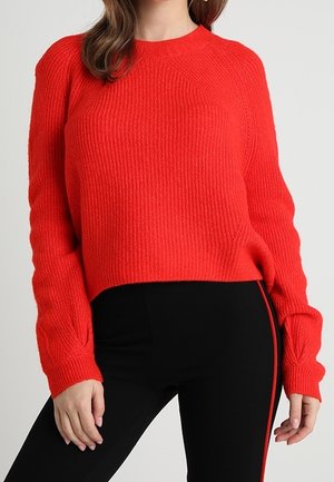 Woman wearing a bright red long-sleeve knit sweater and black pants with a red stripe along the side against a plain background.