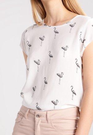Blouse blanche à manches courtes avec petits imprimés de flamants noirs portée avec un pantalon rose clair.