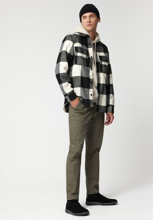 Czarna i biała koszula flanelowa w kratę, z guzikami, beżowy hoodie pod spód, oliwkowe spodnie i czarne buty. Na głowie czarna czapka beanie.