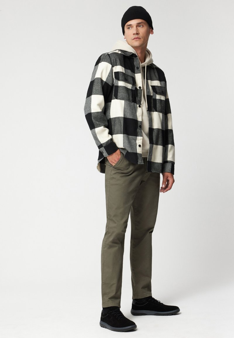 Czarna i biała koszula flanelowa w kratę, z guzikami, beżowy hoodie pod spód, oliwkowe spodnie i czarne buty. Na głowie czarna czapka beanie.