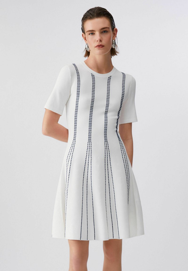 Femme aux cheveux courts portant une robe blanche à manches courtes avec des lignes noires verticales en pointillés, debout devant un fond clair uni.