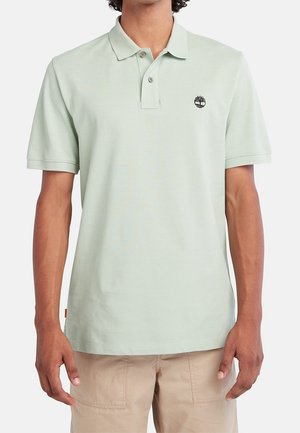Homme portant un polo vert clair à manches courtes avec col et deux boutons, associé à un pantalon beige, visible du cou jusqu’aux hanches.