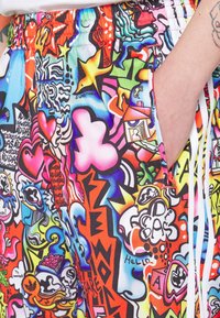 Colorful shorts with vibrant graffiti-style art, including hearts, stars, and various abstract shapes; hand in pocket and visible arm tattoo.