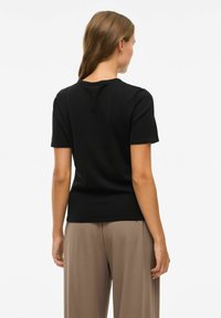 Juoda trumpomis rankovėmis marškinėliai, lygi tekstūra, apykaklė, prigludęs dizainas, dėvimi su kreminės spalvos plačiomis kelnėmis; galinis vaizdas su minimaliais detalėmis.