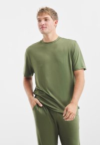 T-shirt verde oliva con maniche corte, realizzato in tessuto morbido. Il modello ha le mani nelle tasche, in piedi contro uno sfondo bianco semplice.