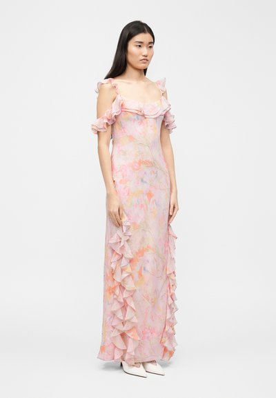 Γυναίκα που φοράει ένα μακρύ, παστέλ φόρεμα με λουλούδια, με βολάν στους ώμους που αφήνουν τους ώμους ακάλυπτους και λεπτομέρειες στο πλάι, σε συνδυασμό με λευκά μυτερά παπούτσια.