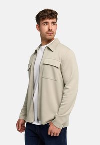 Man wearing light beige zip-up jacket over white shirt and dark jeans, looking to the side against plain background.