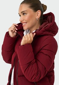 Chaqueta acolchada burdeos con cremallera frontal y capucha acolchada. El material parece ligero, con una textura suave y puños acanalados.
