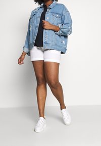 Femme portant une veste en denim déchirée bleu clair, un t-shirt noir, un short blanc et des baskets blanches, se tenant devant un fond blanc uni.