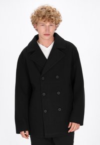 Mustapohjainen villawool peacoat, jossa on kaksirivinen design, ylikokoiset läpät ja suuret mustat napit. Mallilla on päällään valkoinen aluspaita.
