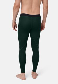 Man standing barefoot facing away, wearing dark green fitted leggings with black waistband on a plain background.