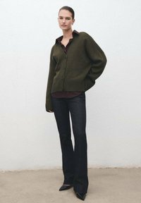 Cárdigan de punto verde oliva con escote en V y cierre de botones, combinado con una camisa oscura y jeans negros acampanados, complementado con tacones puntiagudos.