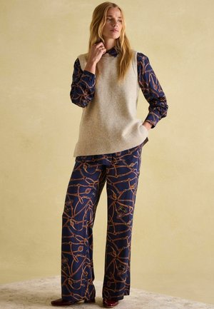 Beige knitted vest over a navy shirt with a chain pattern, paired with wide-leg pants featuring the same design, styled with maroon shoes.