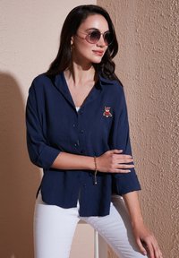 Navy button-up shirt with rolled sleeves, featuring a red and brown teddy bear embroidery. Worn with white pants against a textured wall.
