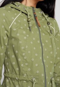 Olive green jacket with small white dot patterns, drawstring hood, zipper closure, elastic cuffs, and adjustable waistband details.