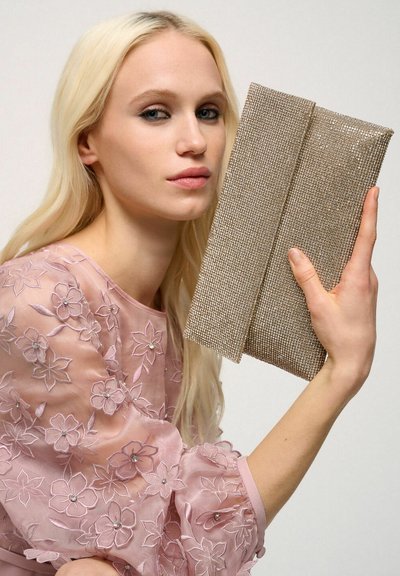 Clutch metalic bej, realizat dintr-un material texturat de plasă, cu un design neted de pliere. Fundalul este neutru, punând în evidență accesoriul.