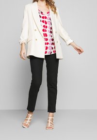 Blazer blanc avec des boutons dorés sur un blouson à motifs roses et rouges, associé à un pantalon noir et des sandales blanches à lanières.