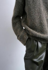 Personne portant un pull marron pelucheux à manches longues et un pantalon en cuir vert foncé, main glissée dans la poche du pantalon, sur un fond uni.