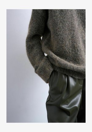 Personne portant un pull marron pelucheux à manches longues et un pantalon en cuir vert foncé, main glissée dans la poche du pantalon, sur un fond uni.