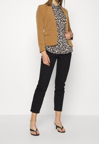 Lichtbruine blazer, overhemd met dierenprint, zwarte slim-fit broek en zwarte sandalen. De blazer heeft zakken; het overhemd heeft een kraag.