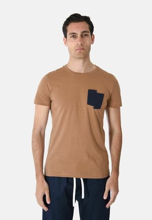 Uomo che indossa una t-shirt marrone a maniche corte con doppia tasca blu navy, abbinata a pantaloni blu navy con coulisse bianchi.