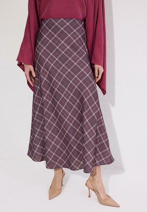 Kobieta ubrana w fioletową kratowaną spódnicę midi w kształcie litery A, bordowy top z długim rękawem oraz beżowe szpilki w szpic, stojąca na jednolitym tle.