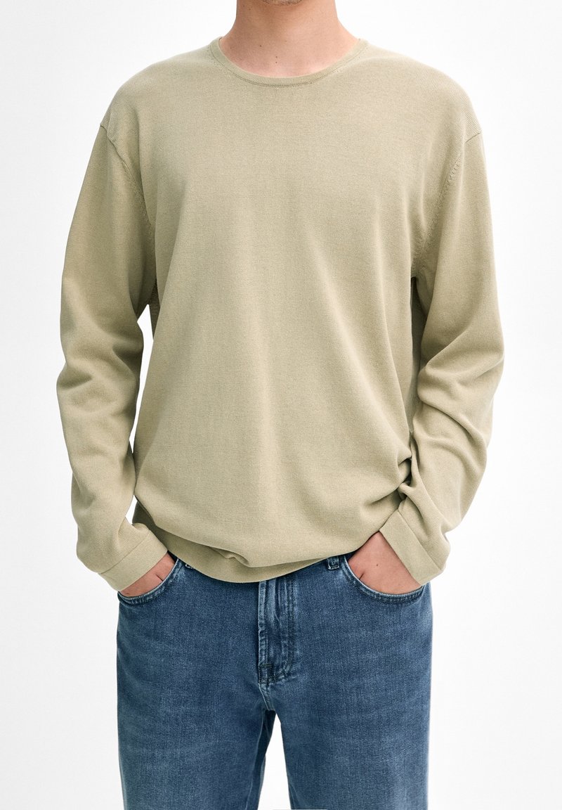 Maglietta a maniche lunghe con scollo tondo in cotone verde oliva chiaro, con vestibilità casual, polsini a coste e una texture morbida. Indossata con jeans di denim blu.