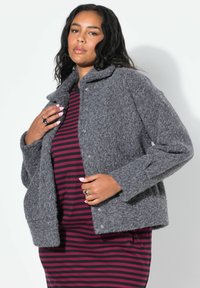 Giacca grigia e pelosa con collo e chiusura a pressione, indossata sopra un vestito a righe bordeaux e nere. Tessuto testurizzato e vestibilità comoda.