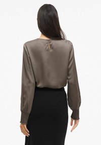 Brun satinbluse med lange ærmer og elastisk smock ved håndleddene. Har en nøglehulslukning på ryggen. Sammen med en sort nederdel.