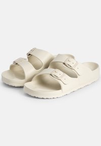 Une paire de sandales à glissière blanc cassé avec deux sangles réglables à boucle et une semelle texturée, conçue pour un usage décontracté.