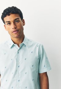 Camicia a maniche corte azzurro chiaro con righe orizzontali e piccoli motivi di fenicotteri ricamati. Presenta un colletto classico e una chiusura con bottoni.