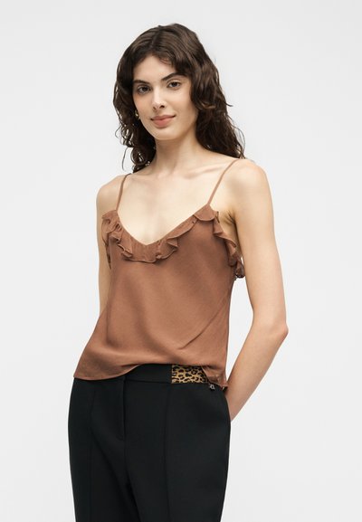 Jeune femme aux cheveux bruns ondulés portant une camisole marron à volants et un pantalon noir avec un détail de ceinture imprimé léopard.