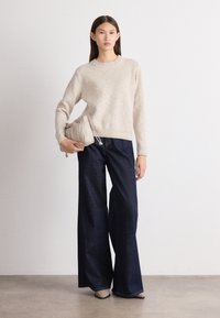 Beige gebreide trui met ribbelrand, gecombineerd met donkere wijde denim jeans. Model houdt een lichtgrijze verzamelde handtas vast.