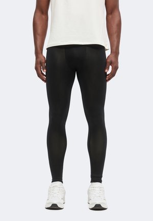 Männliches Model trägt schwarze, enganliegende Leggings und weiße Sneaker, steht vor einem schlichten hellen Hintergrund.