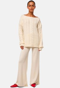 Wollweißer Pullover mit Zopfmuster, weit geschnittenen Ärmeln und gerippten Bündchen, kombiniert mit fließenden cremefarbenen Hosen und roten spitzen Ballerinas.