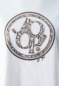 T-shirt blanc avec un graphique rond comportant un "pop" rayé zebra en brun, entouré de petites perles, ajoutant texture et profondeur.