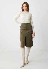 Haut en dentelle blanche à manches longues associé à une jupe midi vert olive avec des fentes sur le devant et des poches, complété par des bottes à talons beiges mi-mollet.