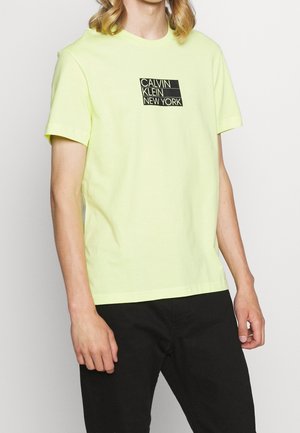 Osoba ubrana w jasnżółtą koszulkę Calvin Klein New York w połączeniu z czarnymi spodniami, stojąca na jednolitym białym tle.